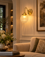 Austin single-light brass wall sconce with a glowing ribbed glass globe installed above a beige sofa in an elegant contemporary living room with warm neutral decor.