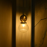 Front view of Austin single-light brass wall sconce glowing warmly at night through its vertically ribbed clear glass globe shade.