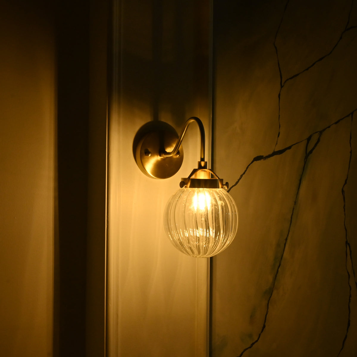 Austin single-light wall sconce shown from the opposite angle, featuring a warm brass finish and glowing ribbed glass globe against a dark interior.