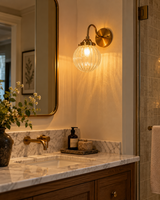 Austin brass ribbed glass globe wall sconce illuminating an elegant bathroom vanity with a marble countertop, gold-framed mirror, and warm ambient lighting.