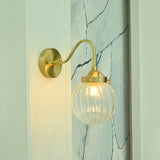 Austin brass wall sconce illuminated with warm light through a clear ribbed glass globe, highlighting its sculptural curved arm and elegant brass detailing.