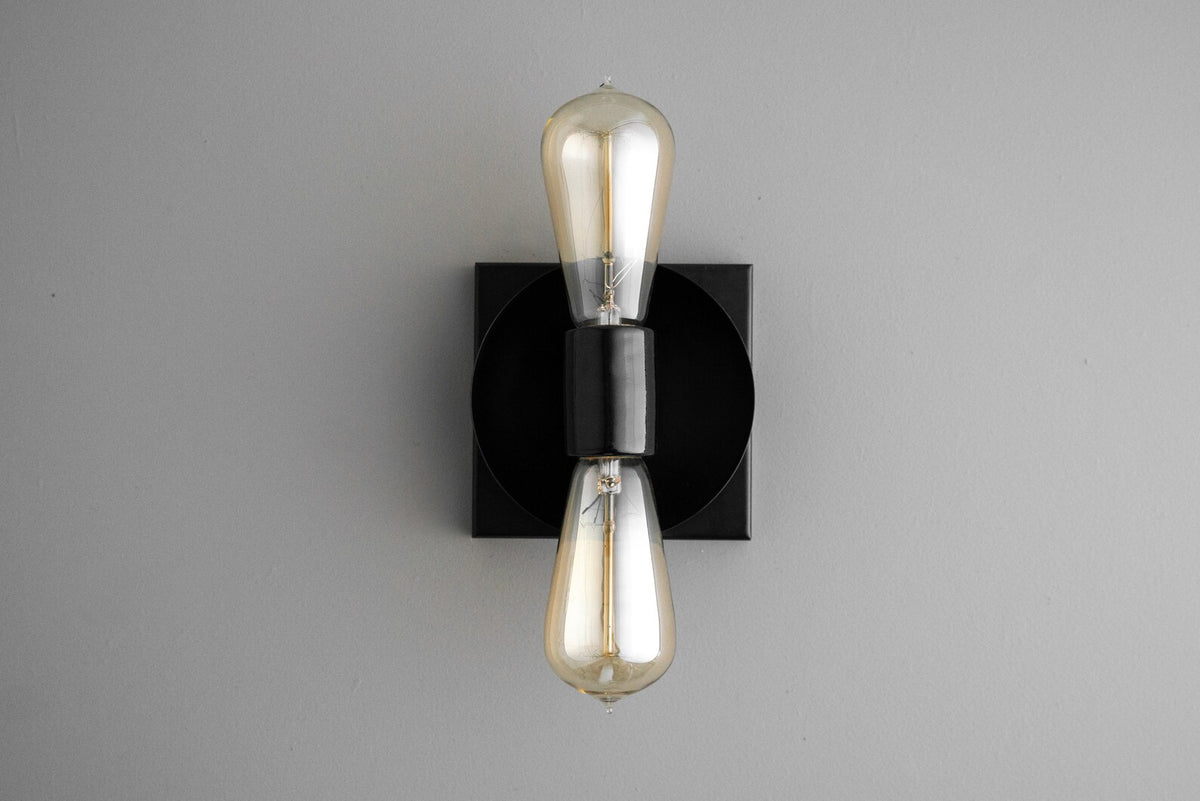 Contemporary wall sconce with black round disc on black square backplate and two vertical Edison-style bulbs, sleek minimalist industrial light.