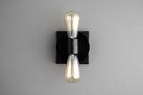 Contemporary wall sconce with black round disc on black square backplate and two vertical Edison-style bulbs, sleek minimalist industrial light.