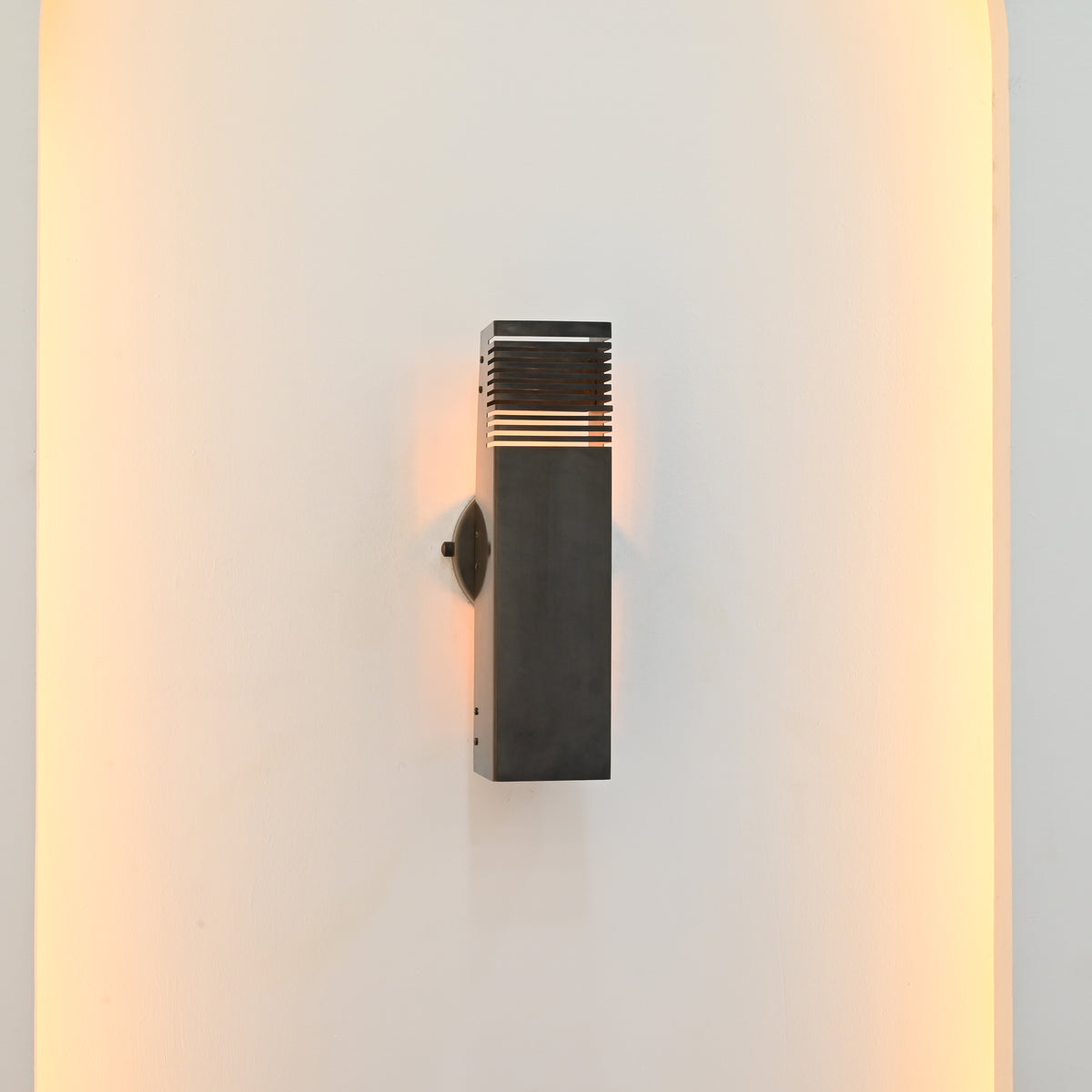 Side angle of Boston 2-light architectural wall sconce highlighting its rectangular metal housing, circular wall backplate, and warm light filtering through horizontal slats.