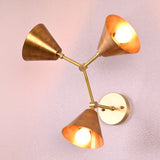 Stylish brass wall-mounted sconce with three conical shades, photographed from a side angle to show depth and structure.