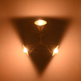 Triple-shade brass wall sconce illuminated in low light, projecting a soft golden glow and triangular light effect on the wall.