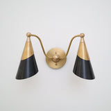 Front view of a two-light brass wall sconce with twin black cone shades and a round brass backplate on a white wall.