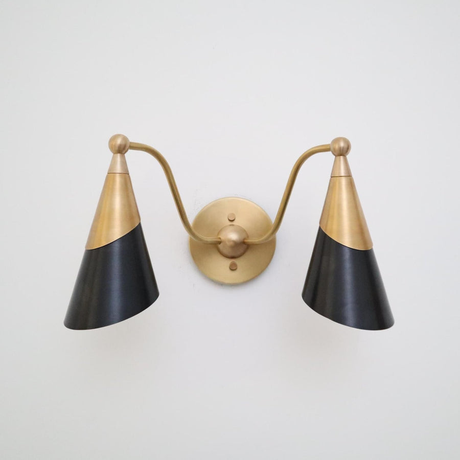 Front view of a two-light brass wall sconce with twin black cone shades and a round brass backplate on a white wall.