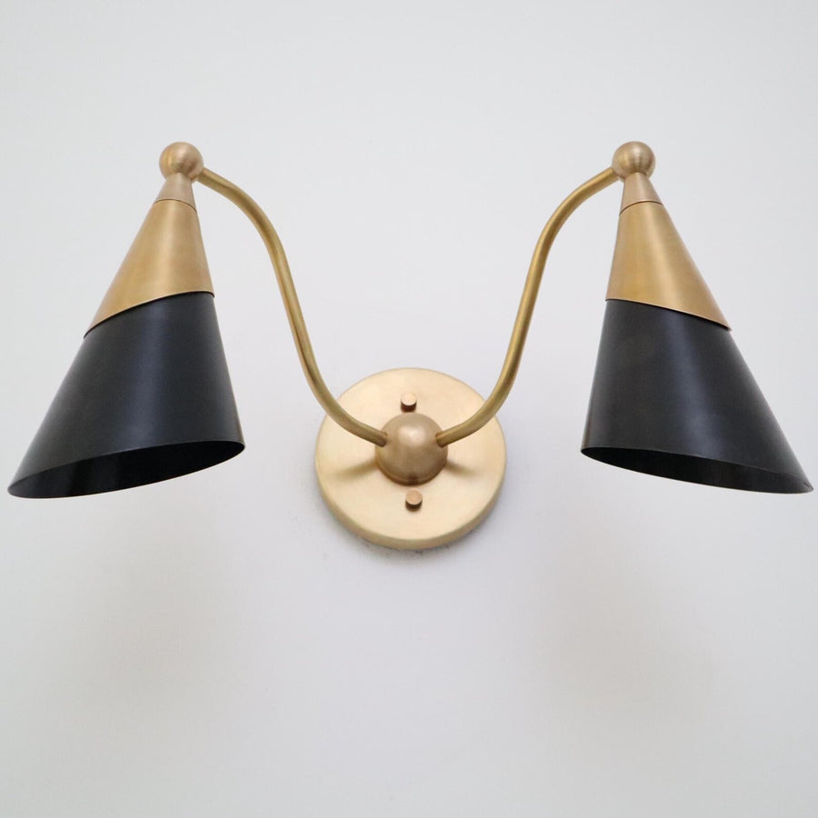Front view of a two-light brass wall sconce with twin black cone shades and a round brass backplate on a white wall.