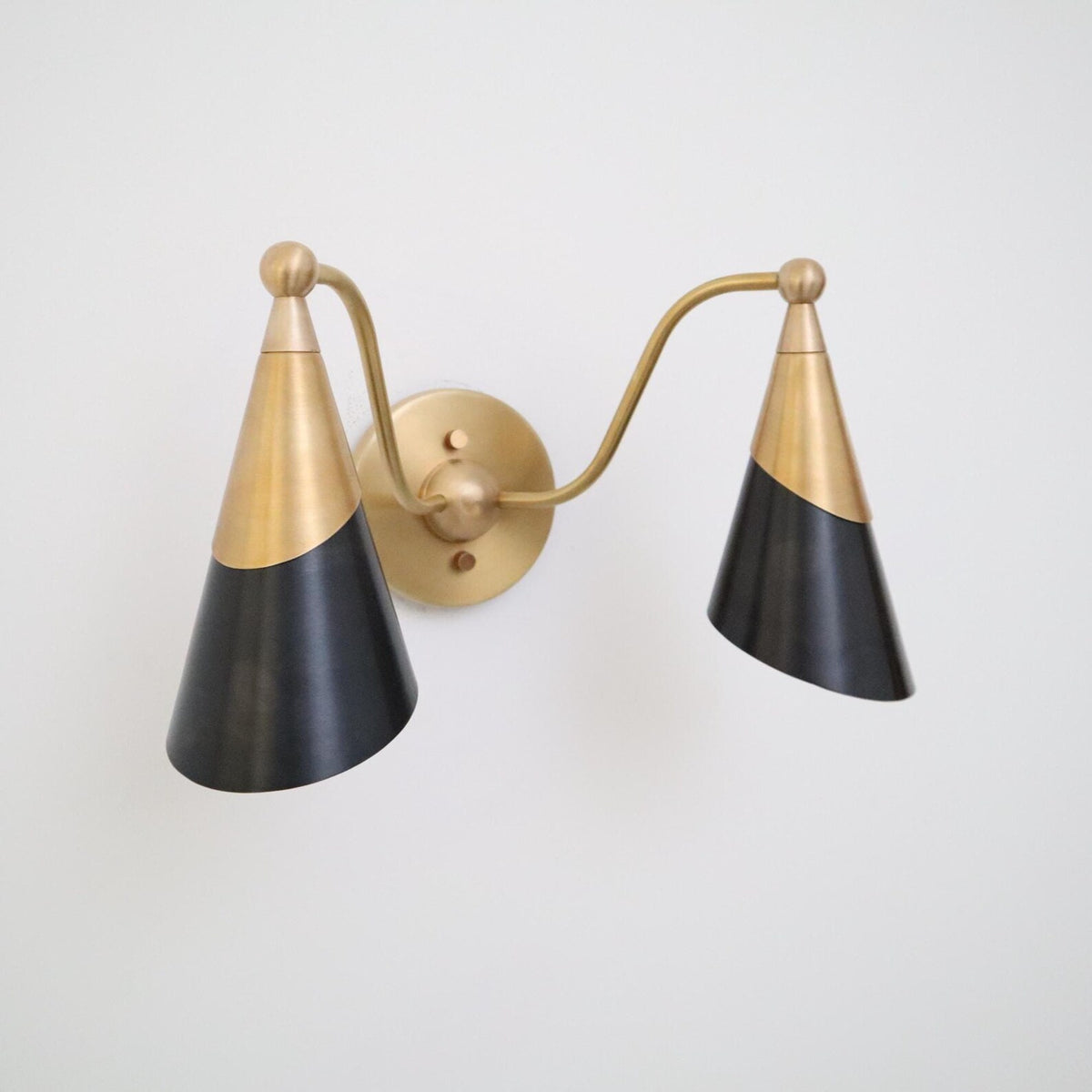 Angled perspective of a modern brass double wall sconce with two-tone black cone shades on a white wall.