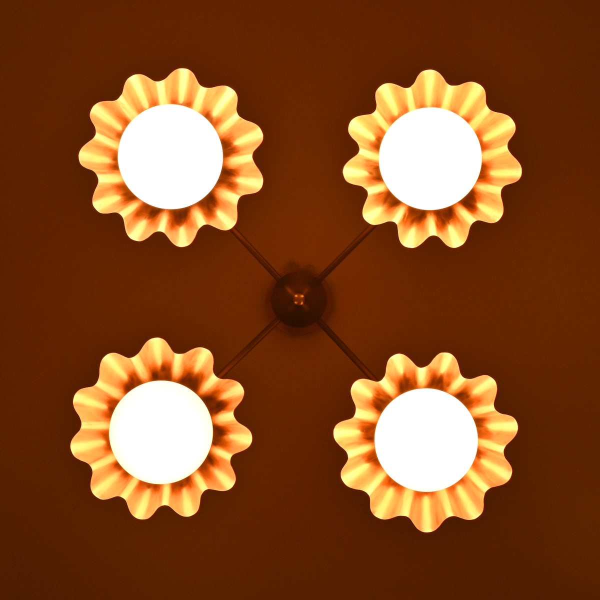 Overhead view of brass chandelier with four glowing ruffled shades, radiating warm golden light in a flower-like pattern.