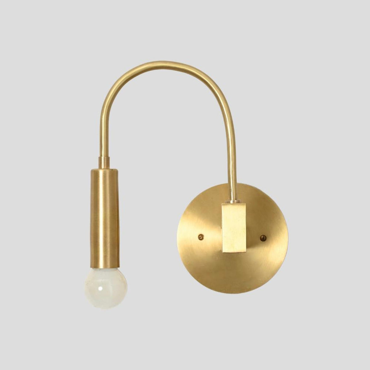 Front-facing view of brass wall sconce with curved arm and minimalist exposed bulb design, mounted on a round brass plate.