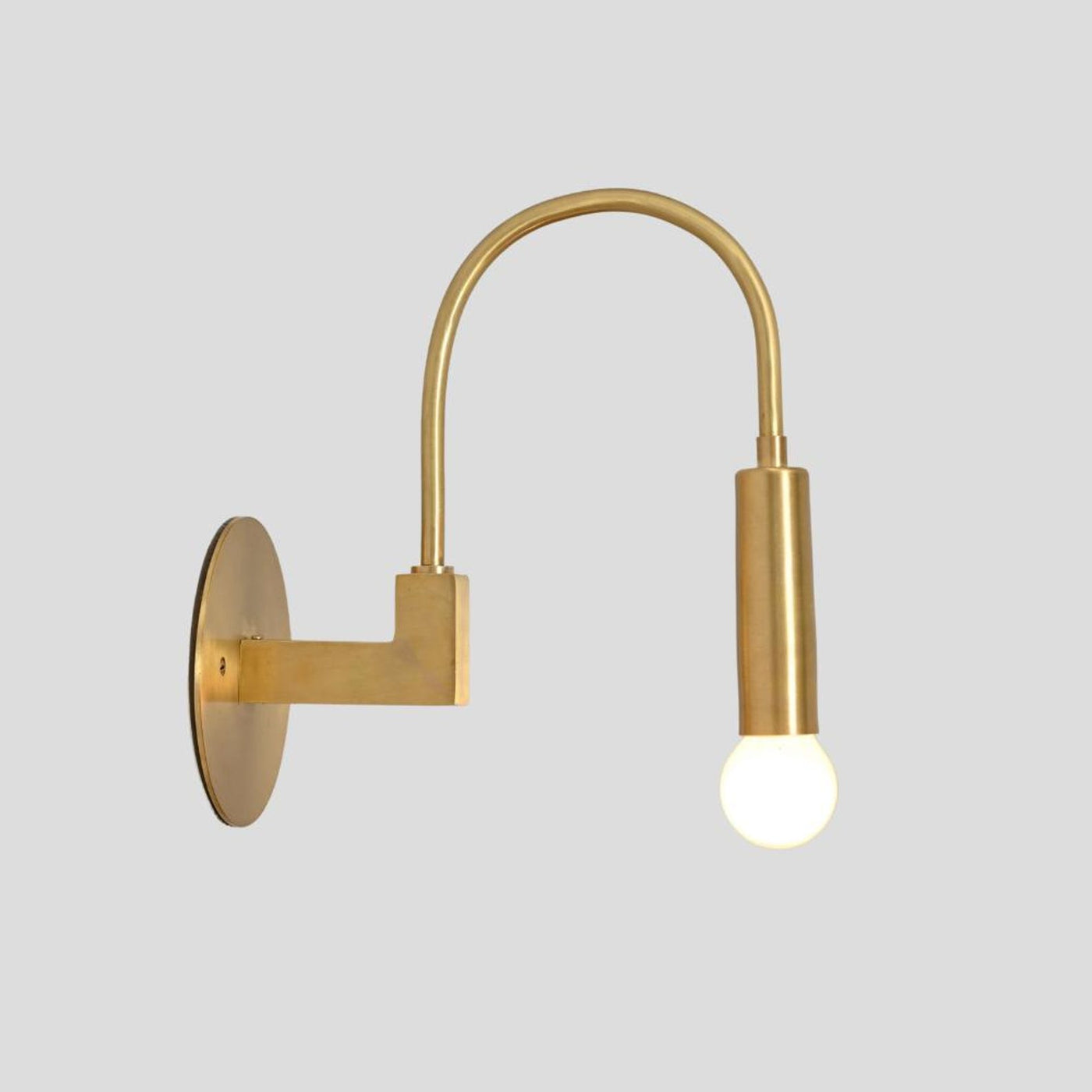 Handmade brass wall sconce with curved arm and exposed bulb, side profile view mounted on round brass base.