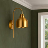 Brass dome wall sconce with curved arm installed on a dark green wall near a window, shown in a styled interior setting.