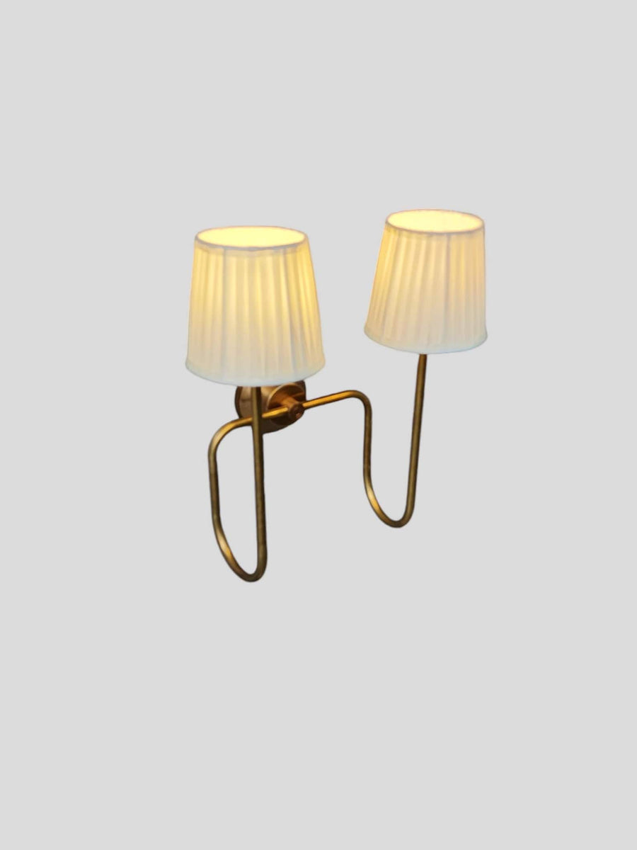 Side angled view of brass double arm wall sconce with pleated white shades, showing curved brass arms and warm ambient glow.