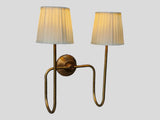 Side angled view of brass double arm wall sconce with pleated white shades, showing curved brass arms and warm ambient glow.