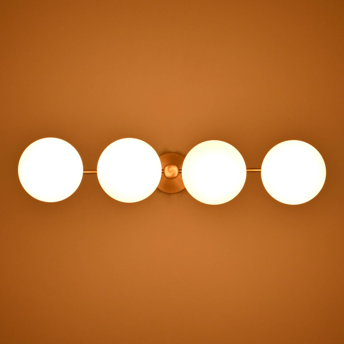 Overhead view of brass chandelier with four glowing frosted globes, radiating soft golden light in a sleek linear design.
