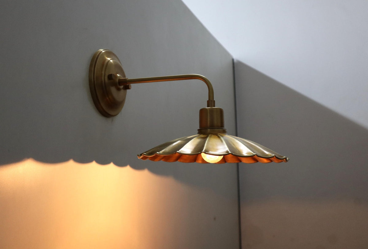 Angled view of a brushed brass scalloped wall sconce with exposed bulb and warm light pattern on the wall.