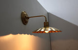 Perspective view of a brass scalloped disc wall sconce showing curved arm, exposed bulb, and warm ambient glow.