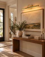 Brisbane brass picture light illuminating framed landscape artwork above a wooden console table in an elegant contemporary entryway.