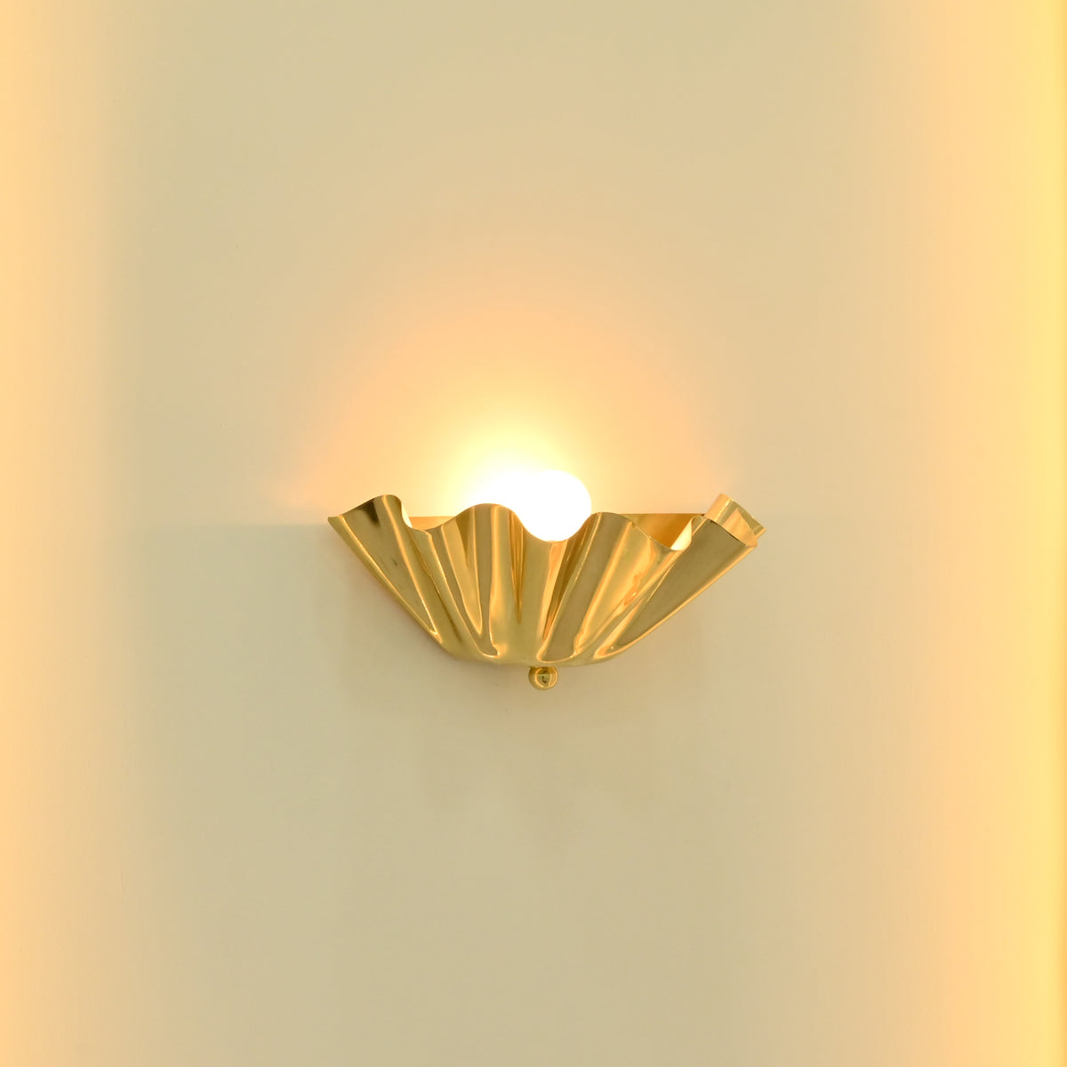 Close-up of Brooklyn single-light brass wall sconce featuring reflective golden folded metal curves and a glowing bulb creating warm uplighting.