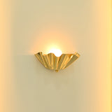 Close-up of Brooklyn single-light brass wall sconce featuring reflective golden folded metal curves and a glowing bulb creating warm uplighting.