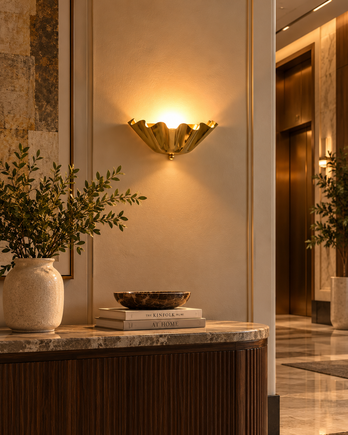 Brooklyn single-light sculptural brass wall sconce casting warm ambient light above a marble-top wooden console in a sophisticated luxury lobby interior.