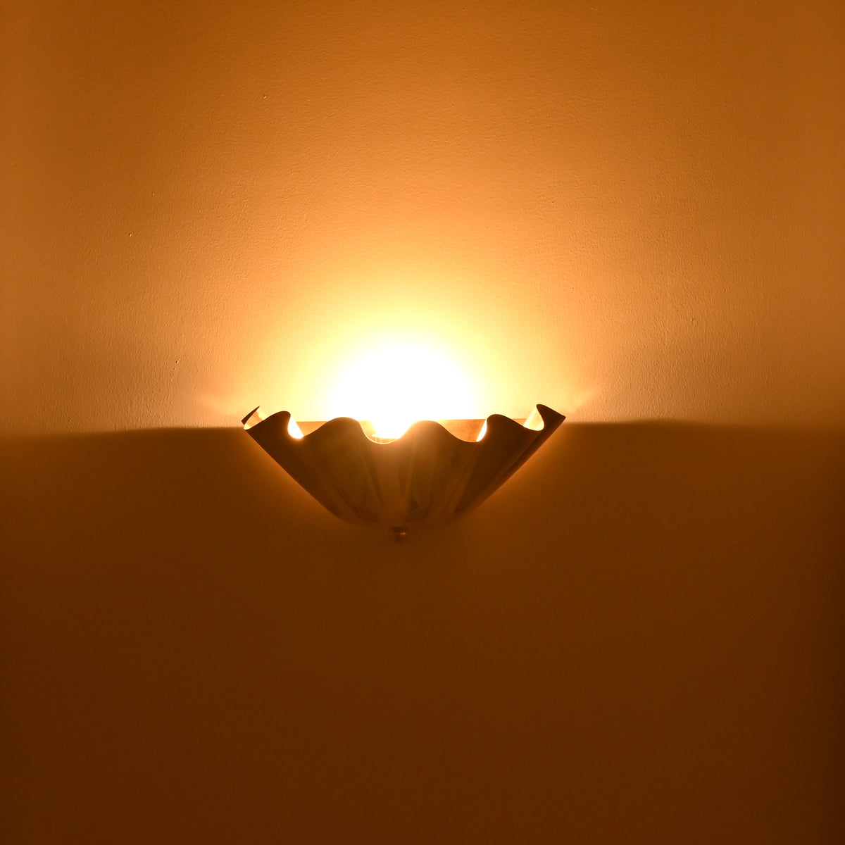 Brooklyn one-light ruffled brass wall sconce creating dramatic upward illumination and a warm ambient lighting effect against a dark golden wall.