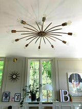 Casablanca 10-light bronze-tone Sputnik chandelier mounted on a living room ceiling, featuring curved metal arms, exposed globe bulbs, decorative starburst rods, and large windows.