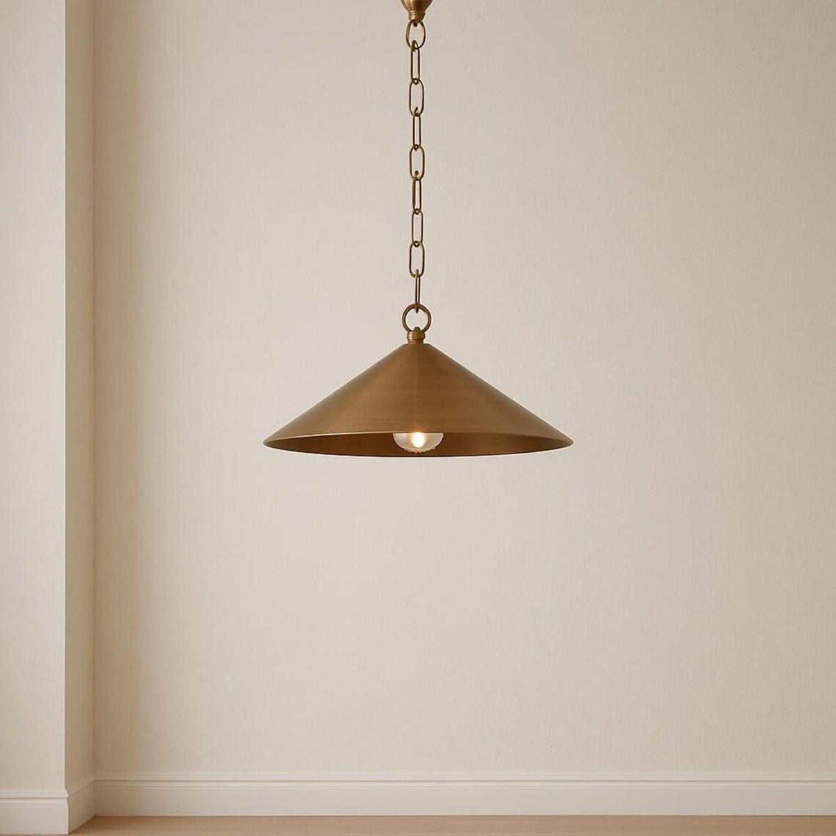 Single brass cone pendant light with chain hanging against a plain beige wall in a minimal interior.