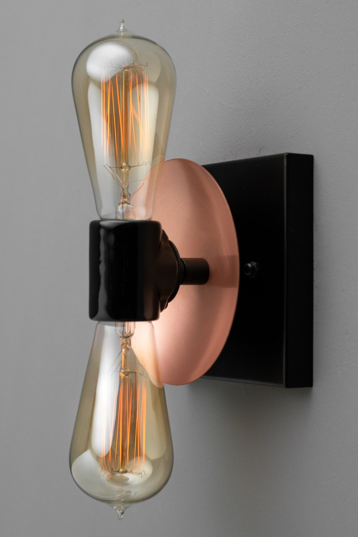 Close-up angled view of wall sconce showing copper disc backplate, black center holder, and glowing Edison bulbs on a gray wall.