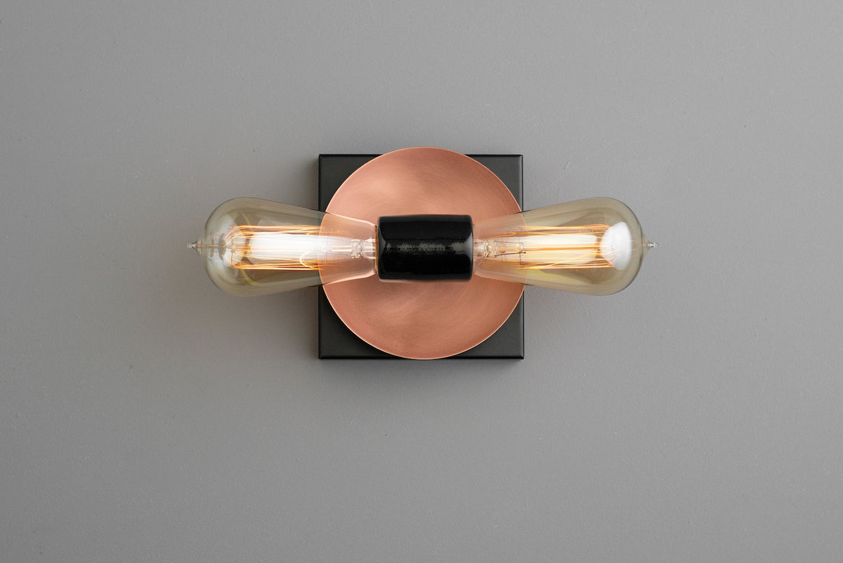 Minimal modern wall light with copper round disc on black square plate and two horizontal filament bulbs, contemporary industrial sconce.