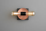 Minimal modern wall light with copper round disc on black square plate and two horizontal filament bulbs, contemporary industrial sconce.