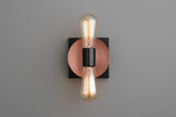 Modern wall sconce with copper disc backplate, black square base, and two vertical Edison-style bulbs, minimalist industrial lighting.