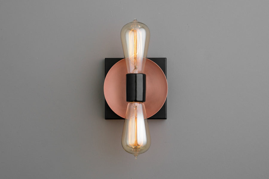 Modern wall sconce with copper disc backplate, black square base, and two vertical Edison-style bulbs, minimalist industrial lighting.