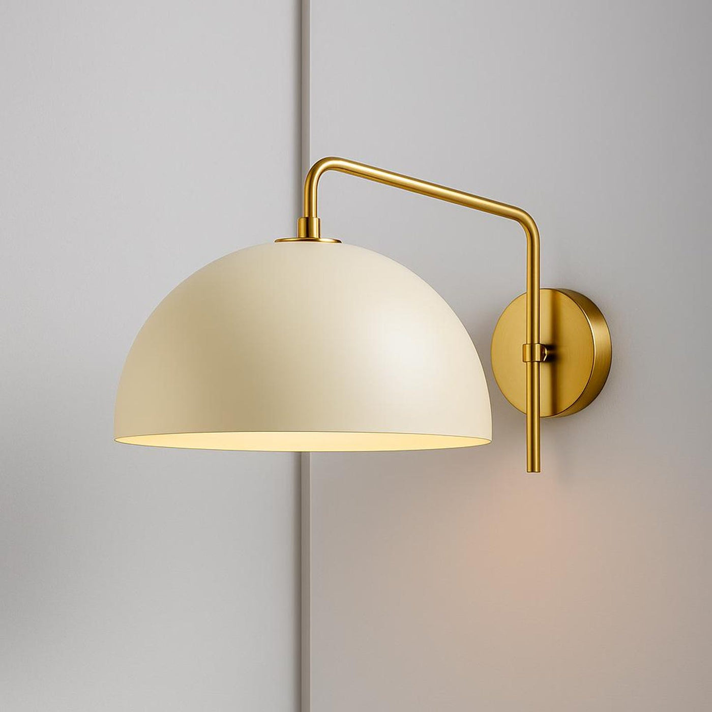 Brushed brass swing-arm dome wall sconce with cream metal shade mounted on a light gray wall.