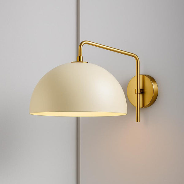 Brushed brass swing-arm dome wall sconce with cream metal shade mounted on a light gray wall.