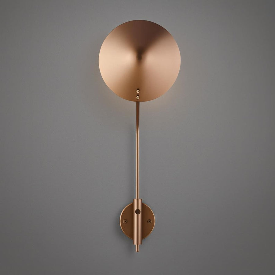 Modern brass disc wall sconce with round shade and curved arm casting a warm halo glow on a light wall.
