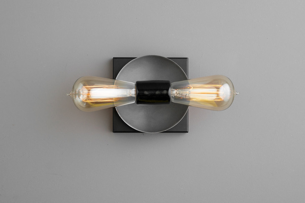 Contemporary wall light with graphite round disc and black square base, twin horizontal Edison bulbs, sleek industrial minimalist design.