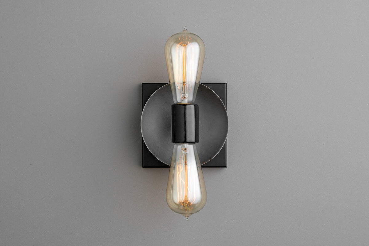 Modern wall sconce with dark graphite round disc on black square backplate and two vertical Edison filament bulbs, minimalist wall lighting.