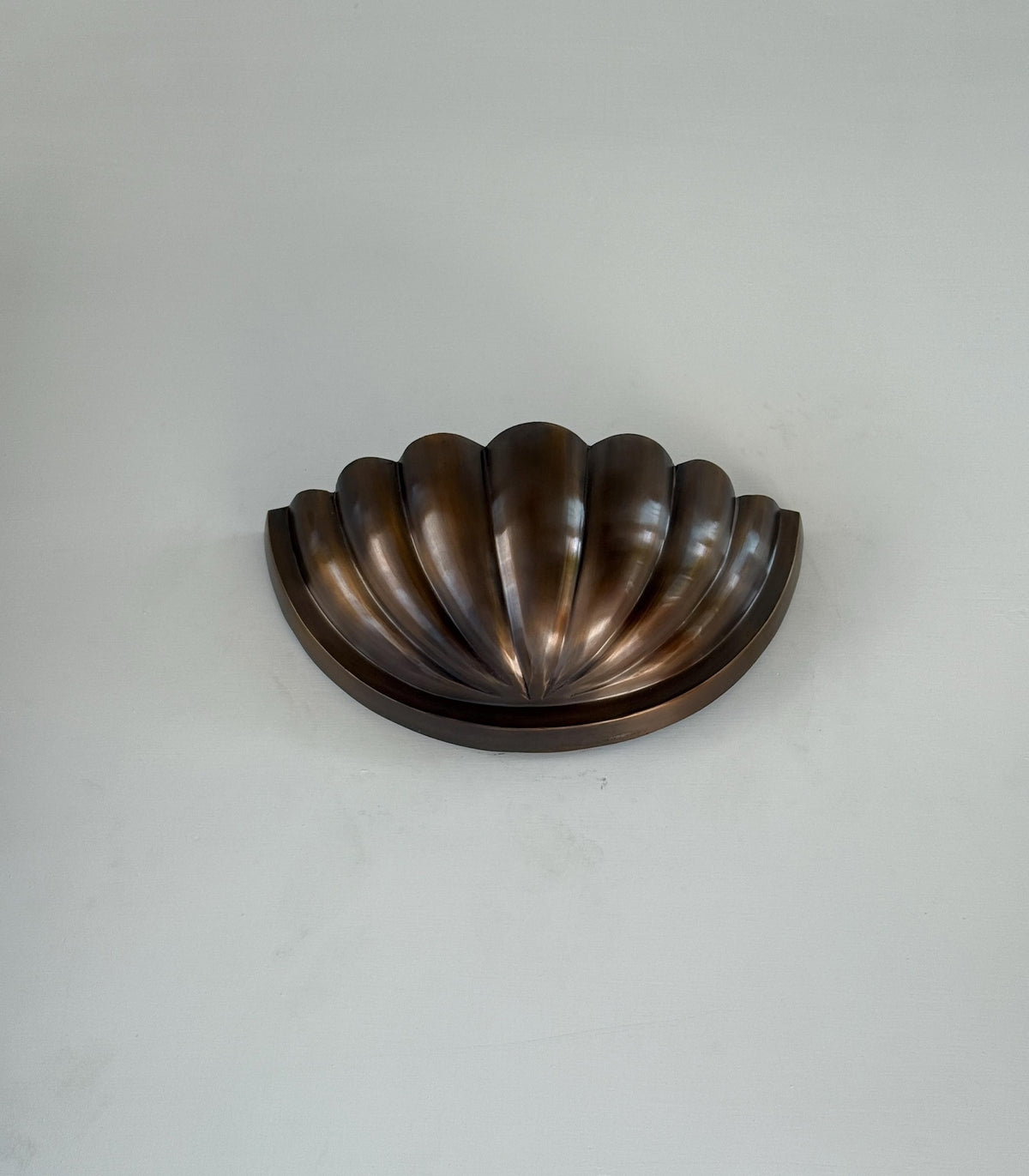 Angled view of fluted scallop brass wall sconce with half-bowl shade in antique bronze finish, mounted on a light wall.