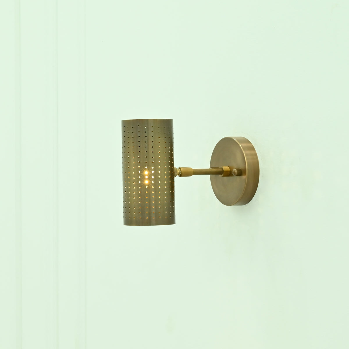 Side view of Hobart perforated brass cylinder wall light with horizontal metal arm, circular backplate, and warm glowing bulb.