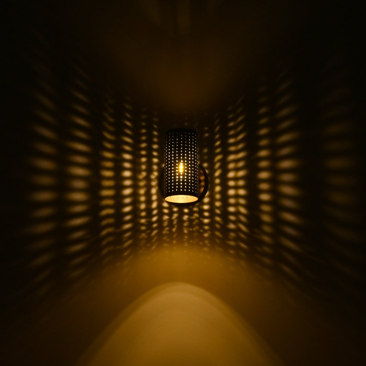 Hobart illuminated perforated cylinder wall sconce casting dramatic warm golden dotted light patterns across a dark wall.