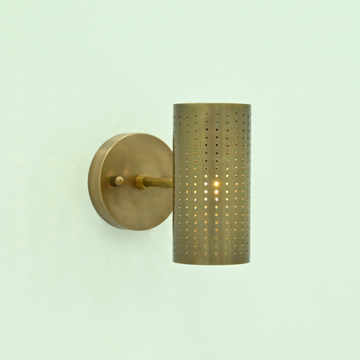  Hobart single-light antique brass wall sconce with illuminated perforated cylindrical metal shade and round mounting backplate.