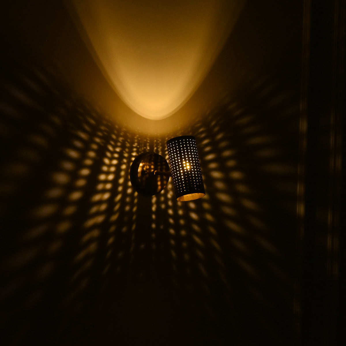 Hobart angled brass cylinder wall sconce projecting golden perforated light patterns and a soft downward beam against a dark wall.