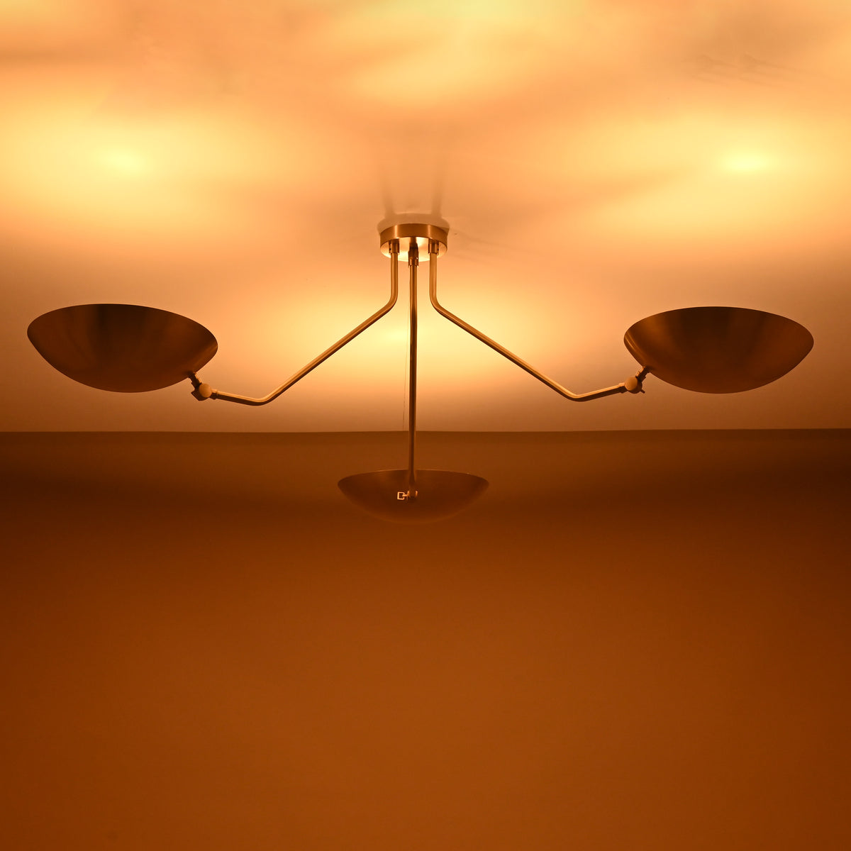 Three-arm brushed brass ceiling light glowing warmly, showing indirect ambient lighting from bowl shades