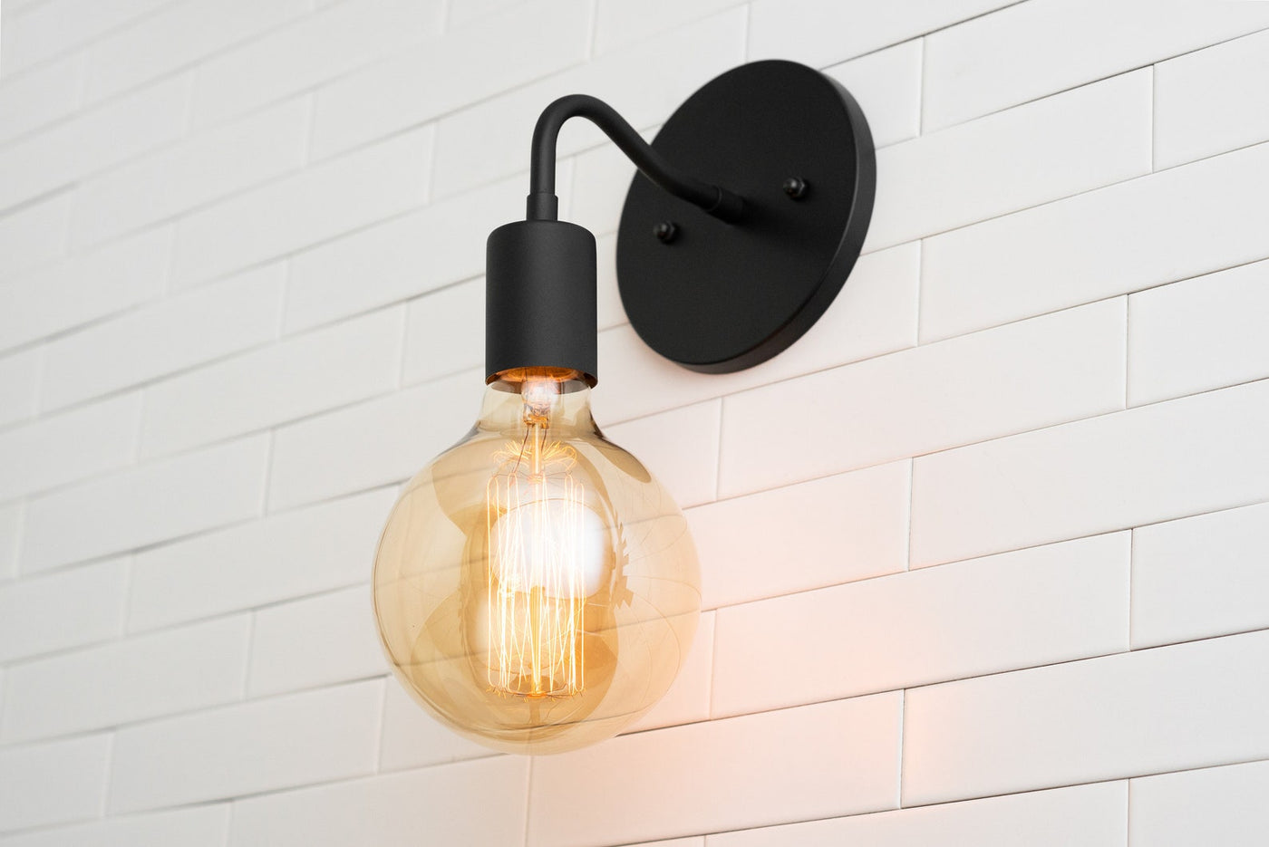 Matte black modern wall sconce with curved arm and warm amber globe Edison bulb on white subway tile wall