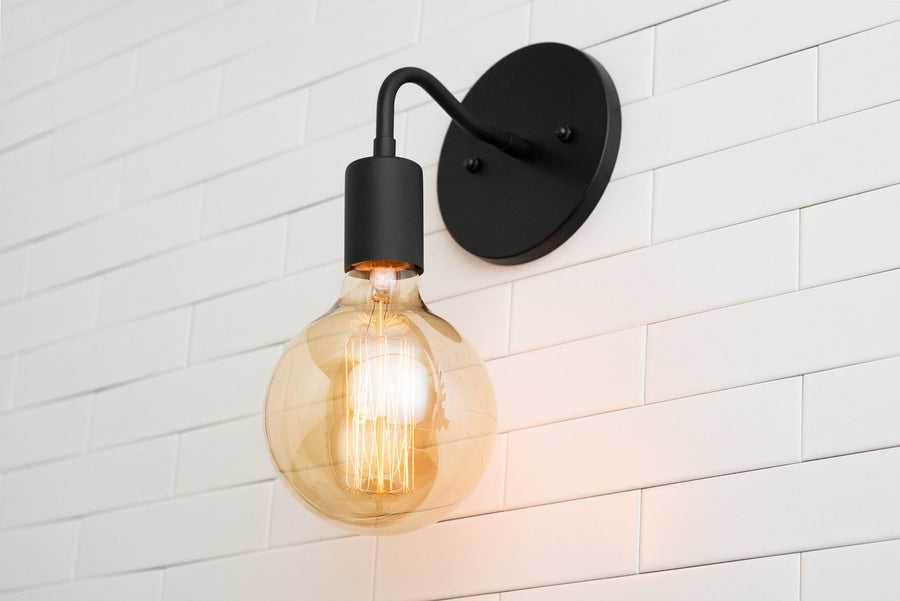 Matte black modern wall sconce with curved arm and warm amber globe Edison bulb on white subway tile wall