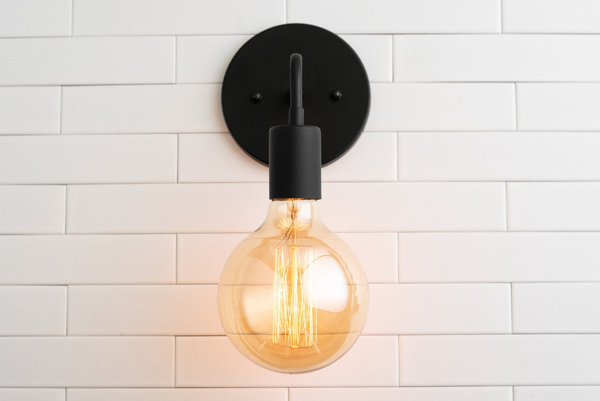 Minimal black wall sconce front view with round backplate and large amber glass globe bulb glowing on white tiled wall