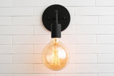 Minimal black wall sconce front view with round backplate and large amber glass globe bulb glowing on white tiled wall